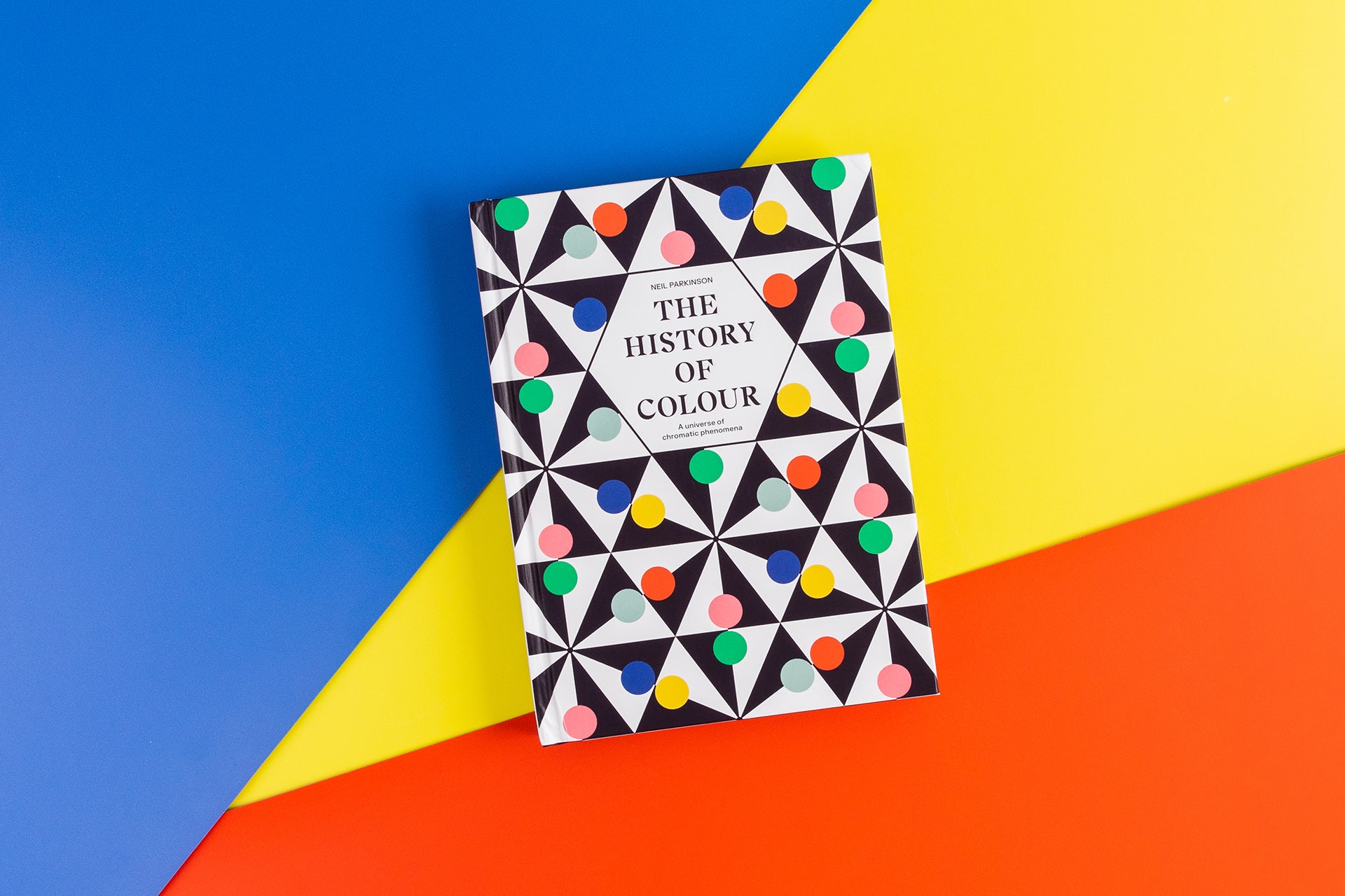 L'image montre la couverture du livre The History of Color, avec une couverture géométrique à pois, disposée sur un fond géométrique bleu, jaune et rouge.