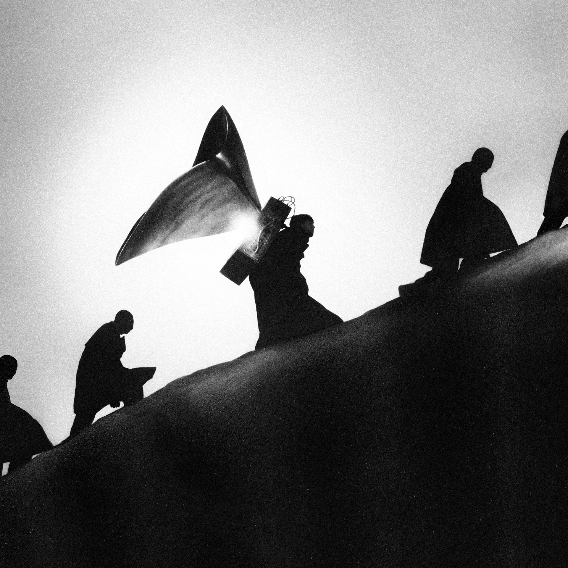 Image en noir et blanc de la pochette de l'album de Playing Robots Into Heaven de James Blake, montrant le musicien portant un grand appareil ressemblant à un tannoy sur le dos, entouré d'autres personnes formant une file d'attente alors qu'ils gravissent une colline qui descend en diagonale sur le tiers inférieur de l'image