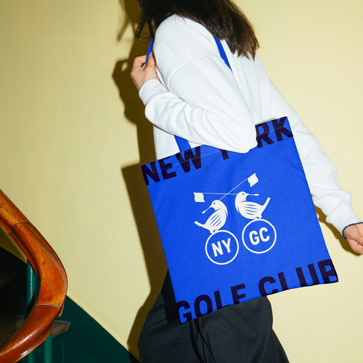 Une personne portant un sac fourre-tout arborant les emblèmes des oiseaux du New York Golf Club tenant un drapeau de golf dans leur bec et les initiales NY GC