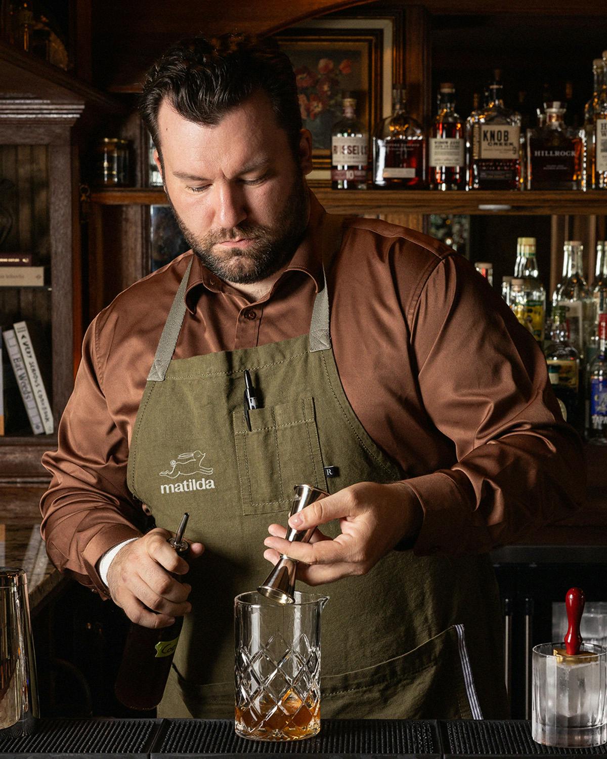 Un barman préparant un cocktail portant une chemise de couleur rouille et un tablier vert sauge avec une illustration de lapin sur la poitrine